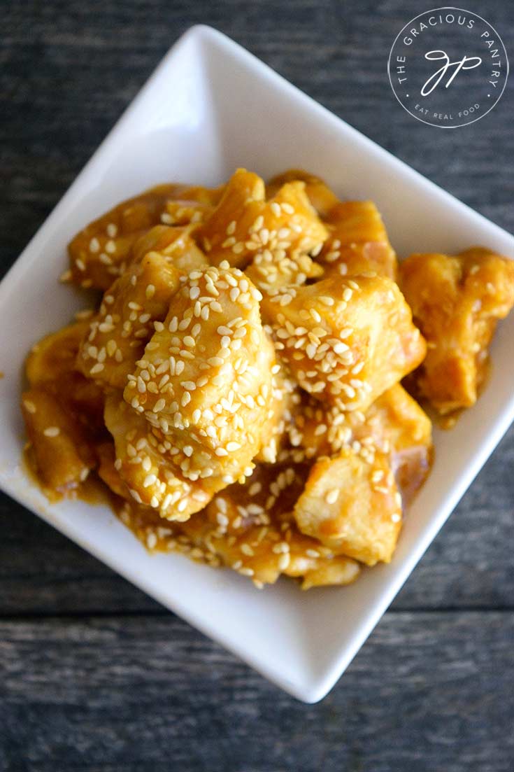 An overhead view looking down into a square, white bowl of this sesame chicken recipe. Sesame seeds are predominantly garnishing the chicken with lots of glaze.