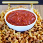 A white bowl of marinara sits on a wooden board surrounded by Air Fryer Pasta Chips.