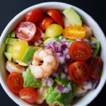 Mixed shrimp and avocado salad in a white bowl on a black background.