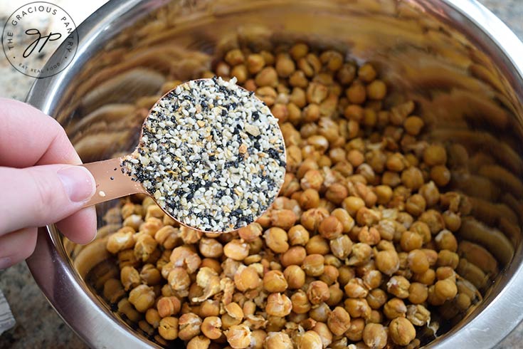 Everything but the bagel seasoning mix being added to a bowl of roasted chickpeas.