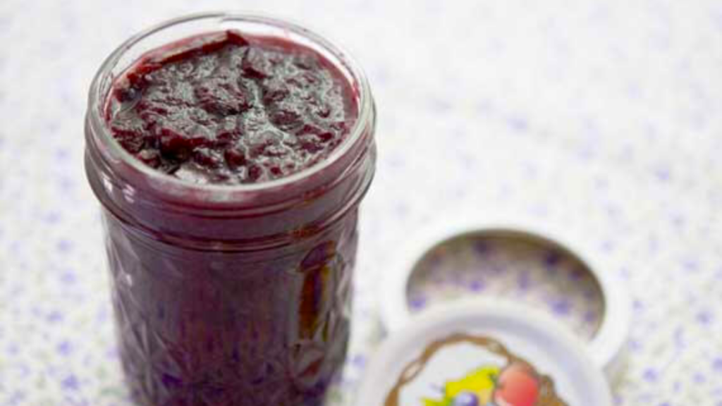 A glass jar sits on a floral cloth and is filled with blueberry apple butter.