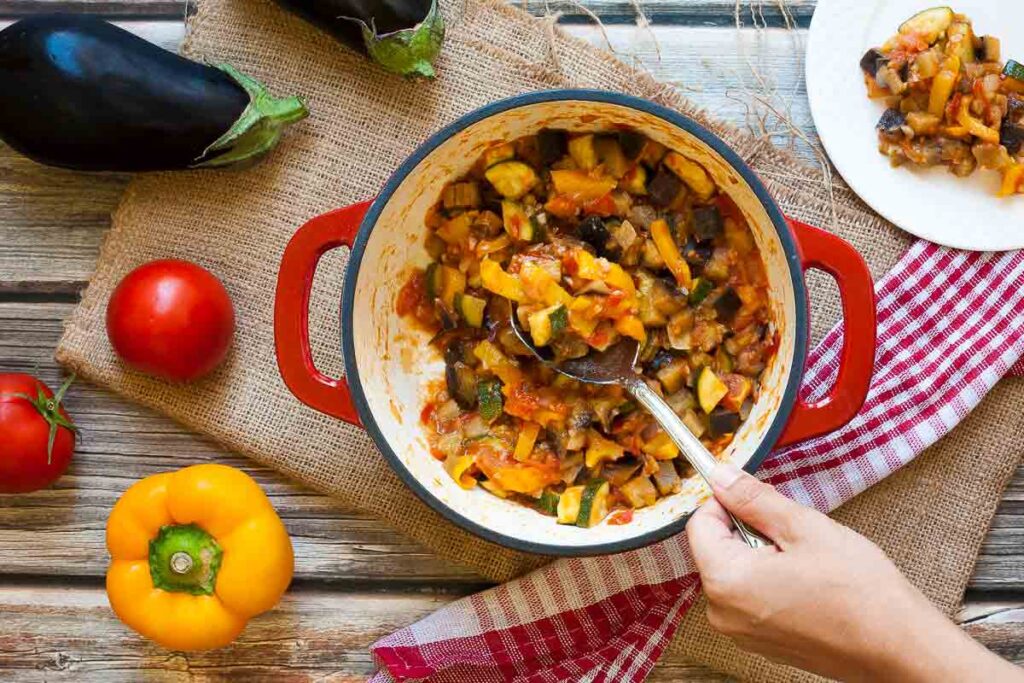 A spoon scoops up a serving of ratatouille from a red pot.