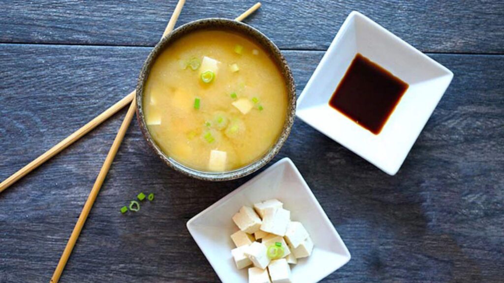 An overhead view of a bowl of miso soup next to two white bowls holding soy sauce and tofu cubes. Chopsticks sit to the side of the bowl.