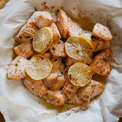 An overhead view of a parchment-lined basket filled with air fryer salmon bites and topped with thin lemon slices.
