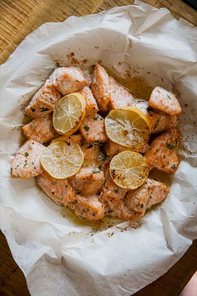 An overhead view of a parchment-lined basket filled with air fryer salmon bites and topped with thin lemon slices.