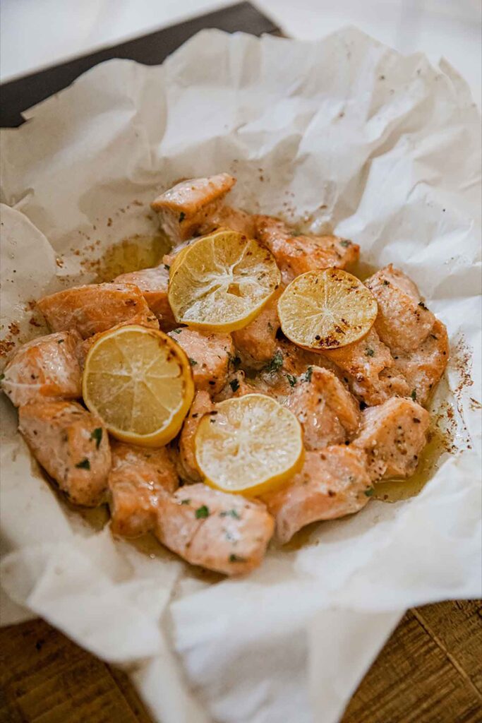 A side view of a parchment-lined basket filled with air fryer salmon bites and topped with lemon slices.