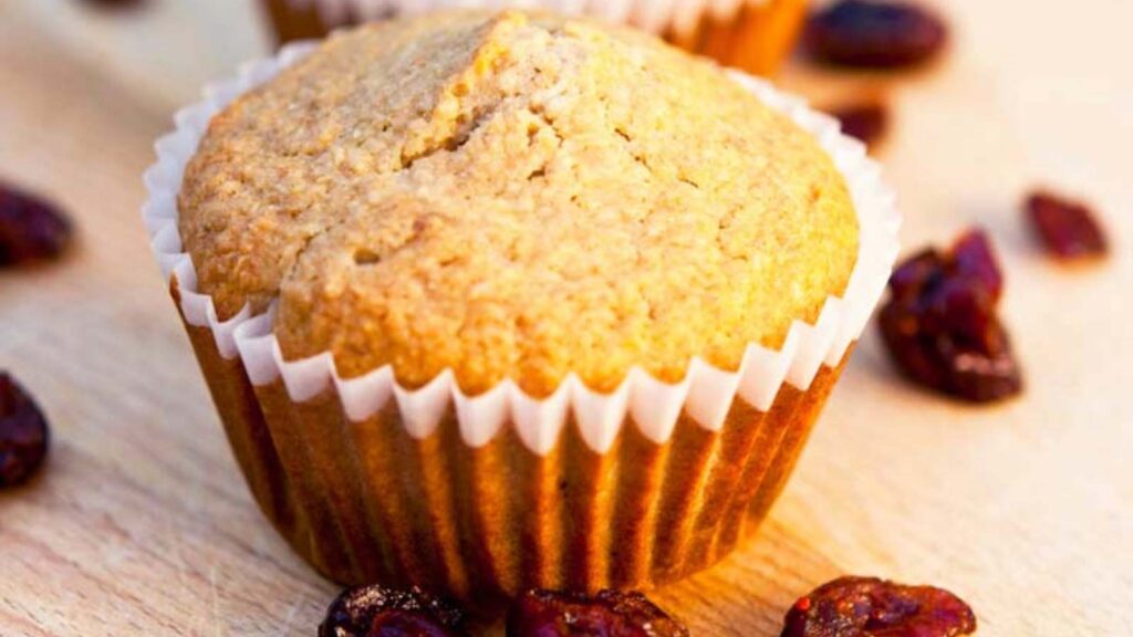 A closeup of a Cranberry Oat Bran Muffin with dried cranberries laying around it on a wood surface.