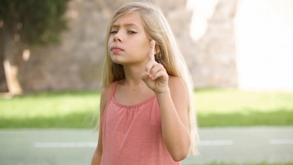 A little girl with blond hair holds up her index finger with a scowl on her face.