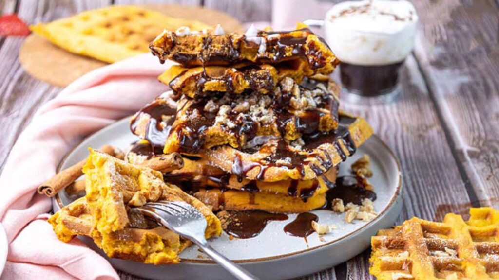 A large stack of pumpkin waffles served on a plate with chocolate syrup and whipped cream over the top.