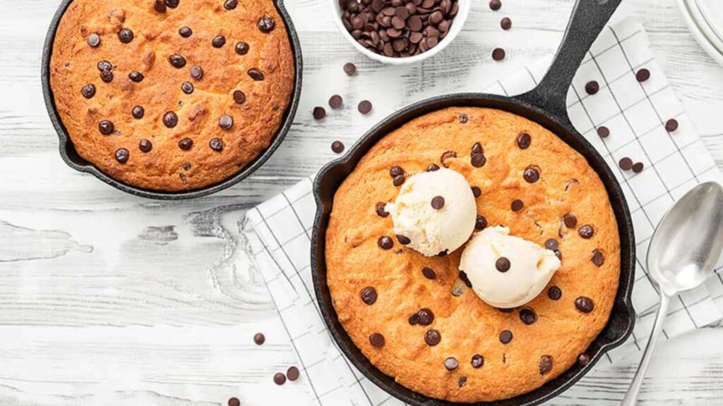 Two skillet chocolate chips cookies in cast iron pans. One has two scoops of vanilla on top.