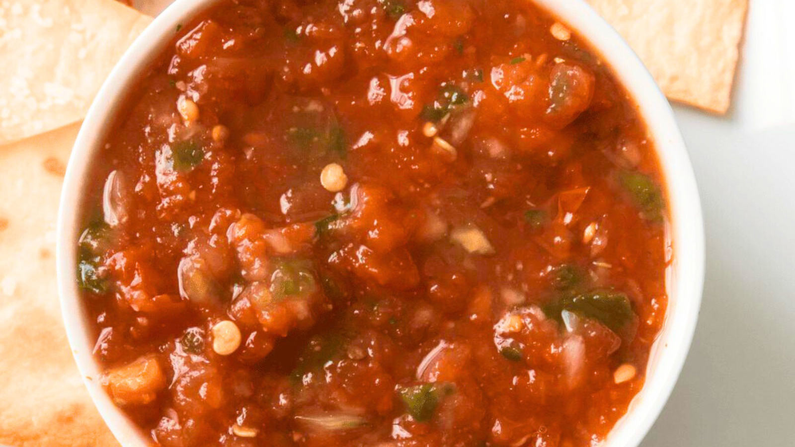 An overhead view of salsa in a white bowl with a few chips around it.