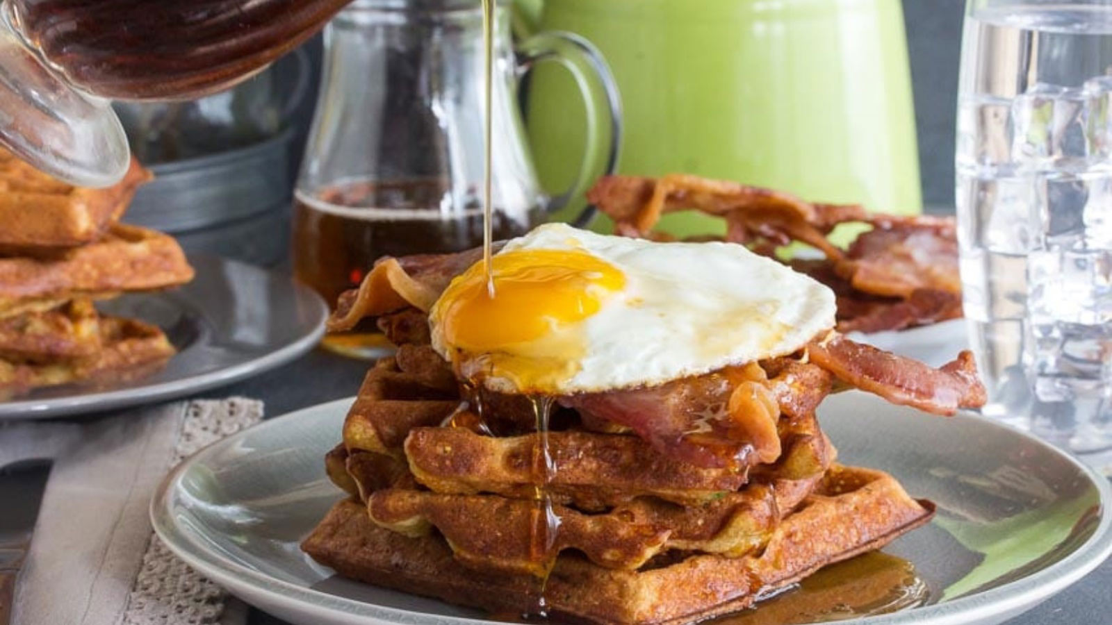 A stack of cheddar waffles with bacon. A fried egg lays on top and maple syrup is being poured over the top.