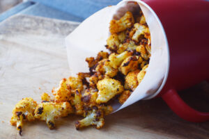 A red mug, tipped over, with Cauliflower Popcorn cascading out of it onto a wood cutting board.