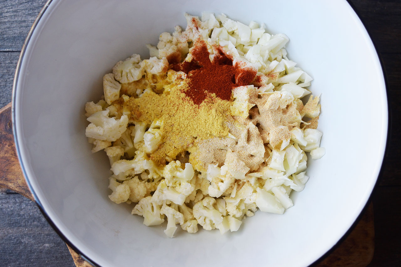 Spices and oil added to raw cauliflower bits in a white mixing bowl.