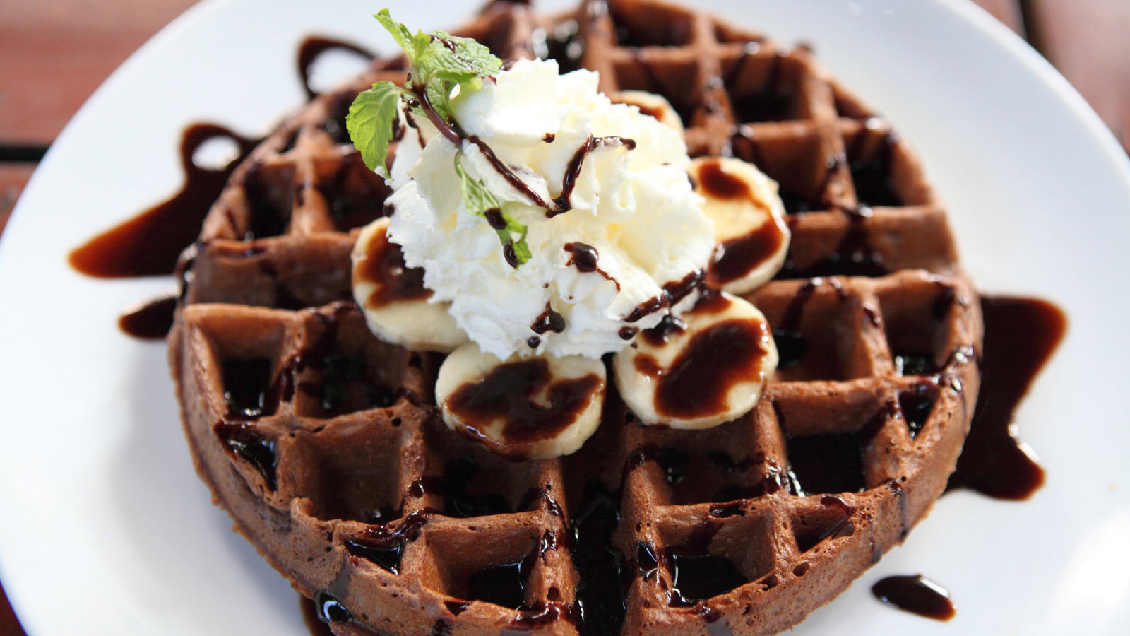 A single chocolate waffle with whipped cream and chocolate drizzle on a white plate.