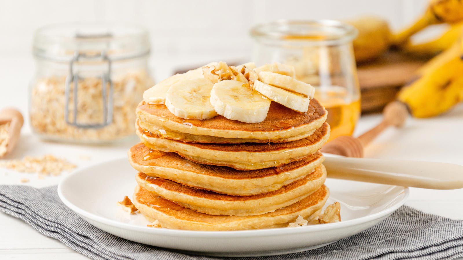 A stack of banana pancakes with slices of fresh bananas, walnuts and honey on top with cup of tea on a white wooden background. A healthy breakfast.