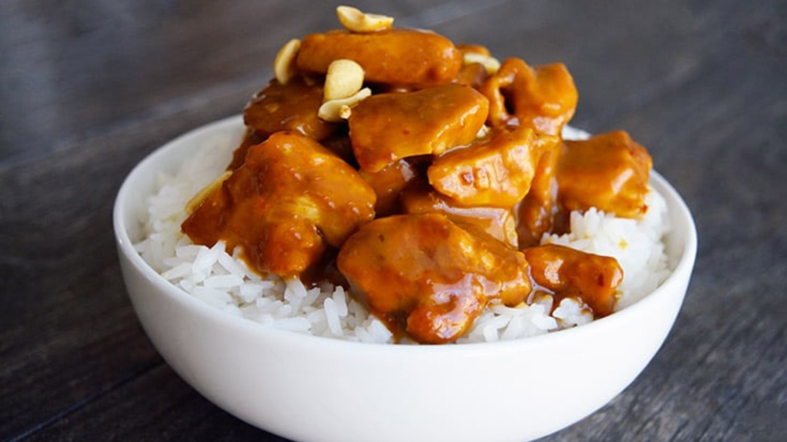 A white bowl with a base of rice and peanut butter chicken piled high on top and garnished with peanuts.