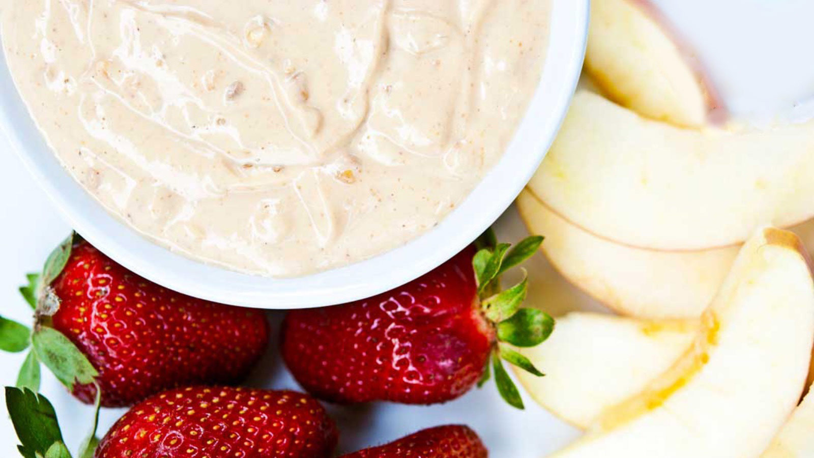 An overhead view of a white bowl filled with peanut butter fruit dip. Fresh strawberries and apple slices lay around the bowl.