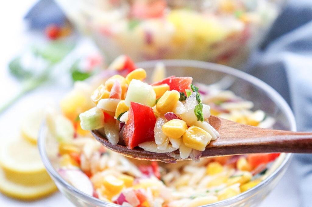 A wooden spoon lift up a spoonful of Rainbow Orzo Salad from a bowl.