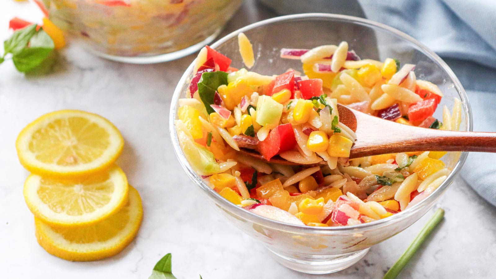 A glass bowl filled with Rainbow Orzo Salad with a wooden spoon resting in it.