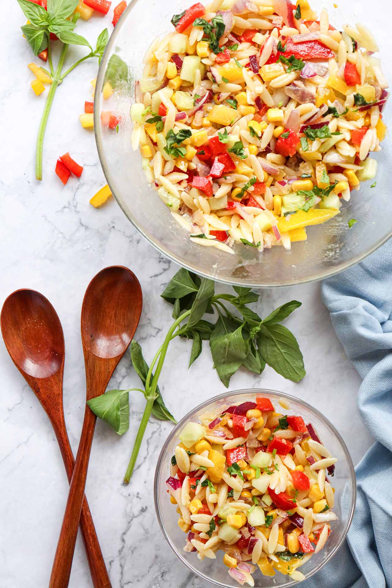 An overhead view of two glass bowls filled with rainbow orzo salad, sitting on a white table with two wooden spoons.