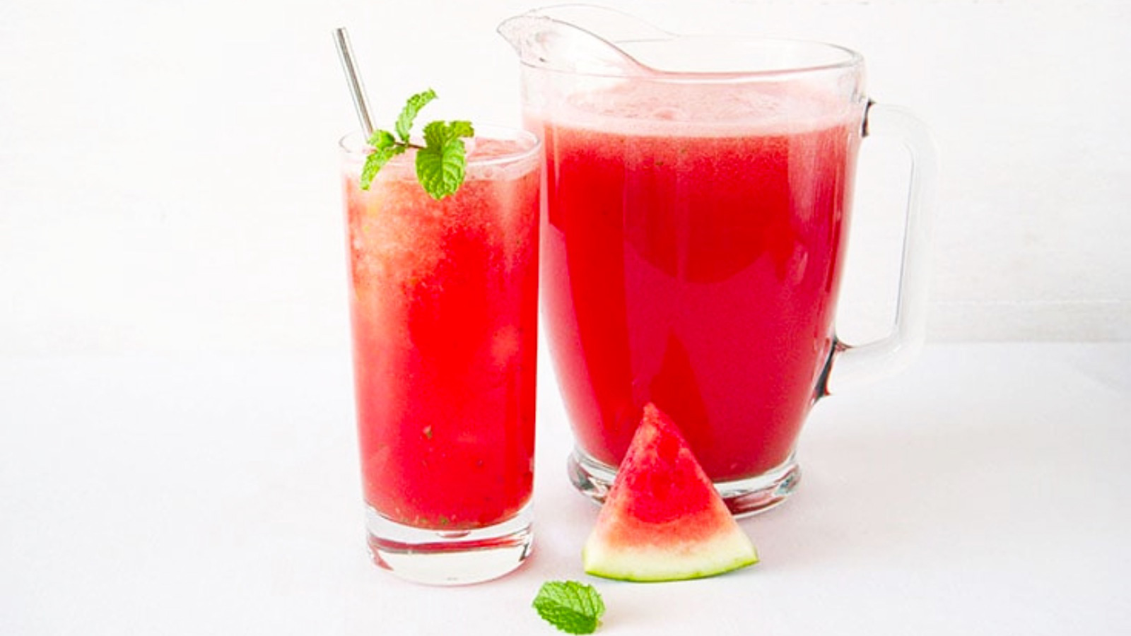 A glass and pitcher on a white surface both sit full of watermelon aqua fresca. The glass is garnished with a fresh mint sprig and a metal straw.