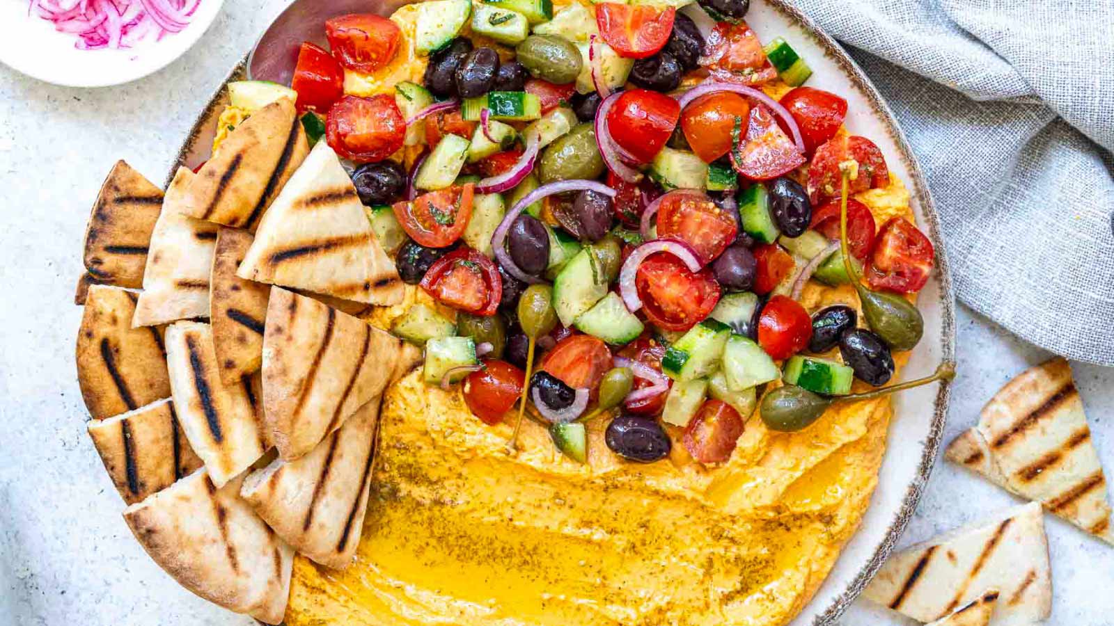 An overhead view of a bowl of hummus loaded with fresh toppings like tomatoes, olives, cucumbers, and pita chips.