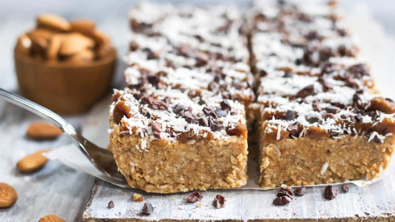 Magic bars lined up on a wood surface, cut into serving sizes. A spatula reaches under one bar to lift it up.