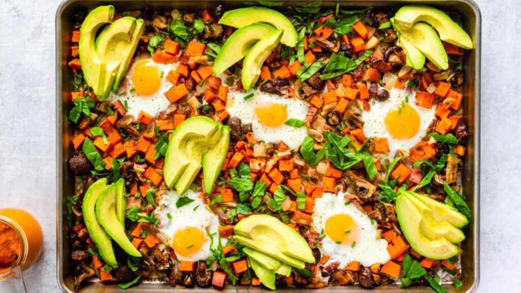 An overhead view of a sheet pan filled with sweet potato hash, baked eggs and avocado slices.