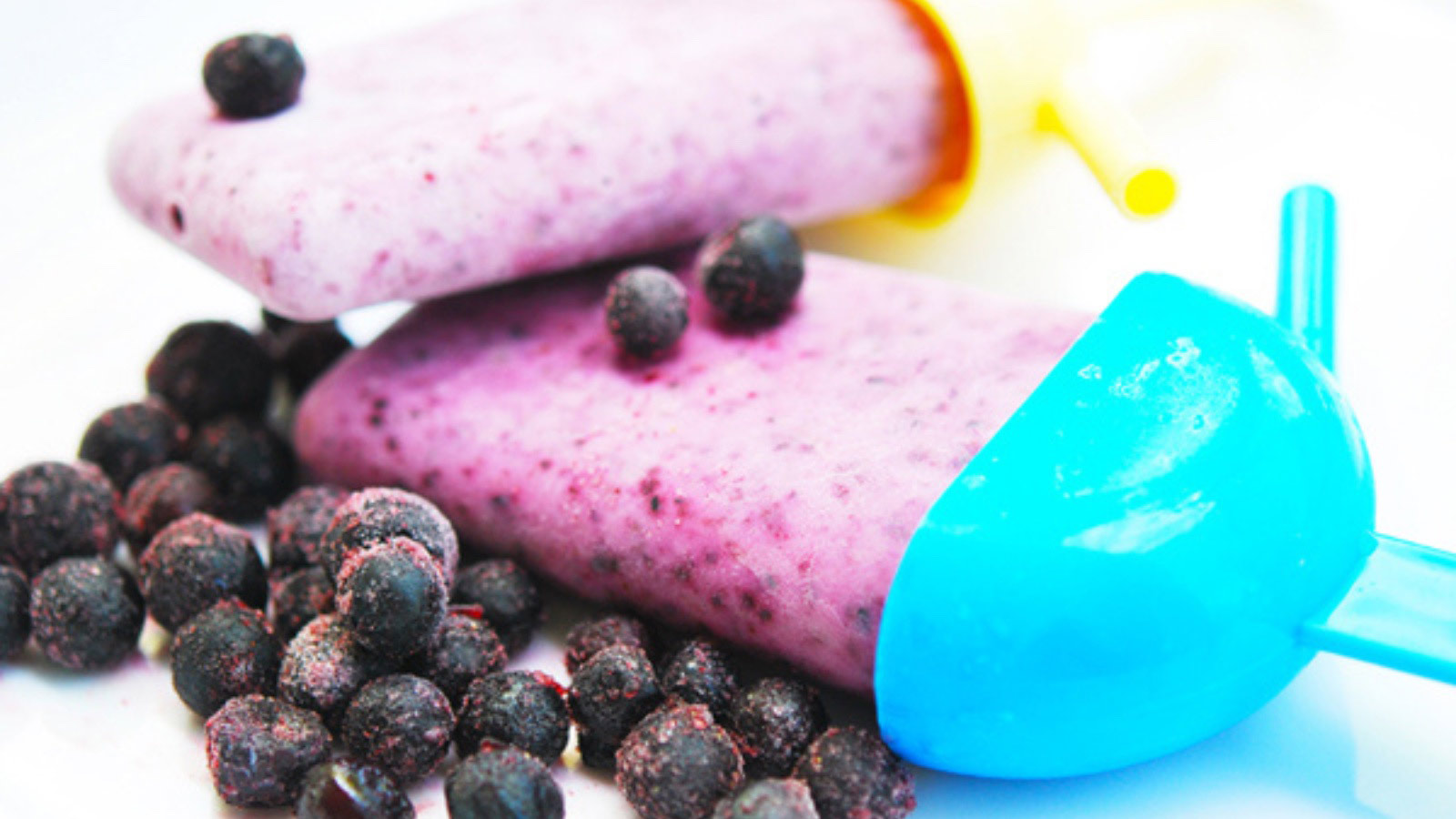 Two Blueberry Yogurt Popsicles laying on a white background with frozen blueberries in front of them.
