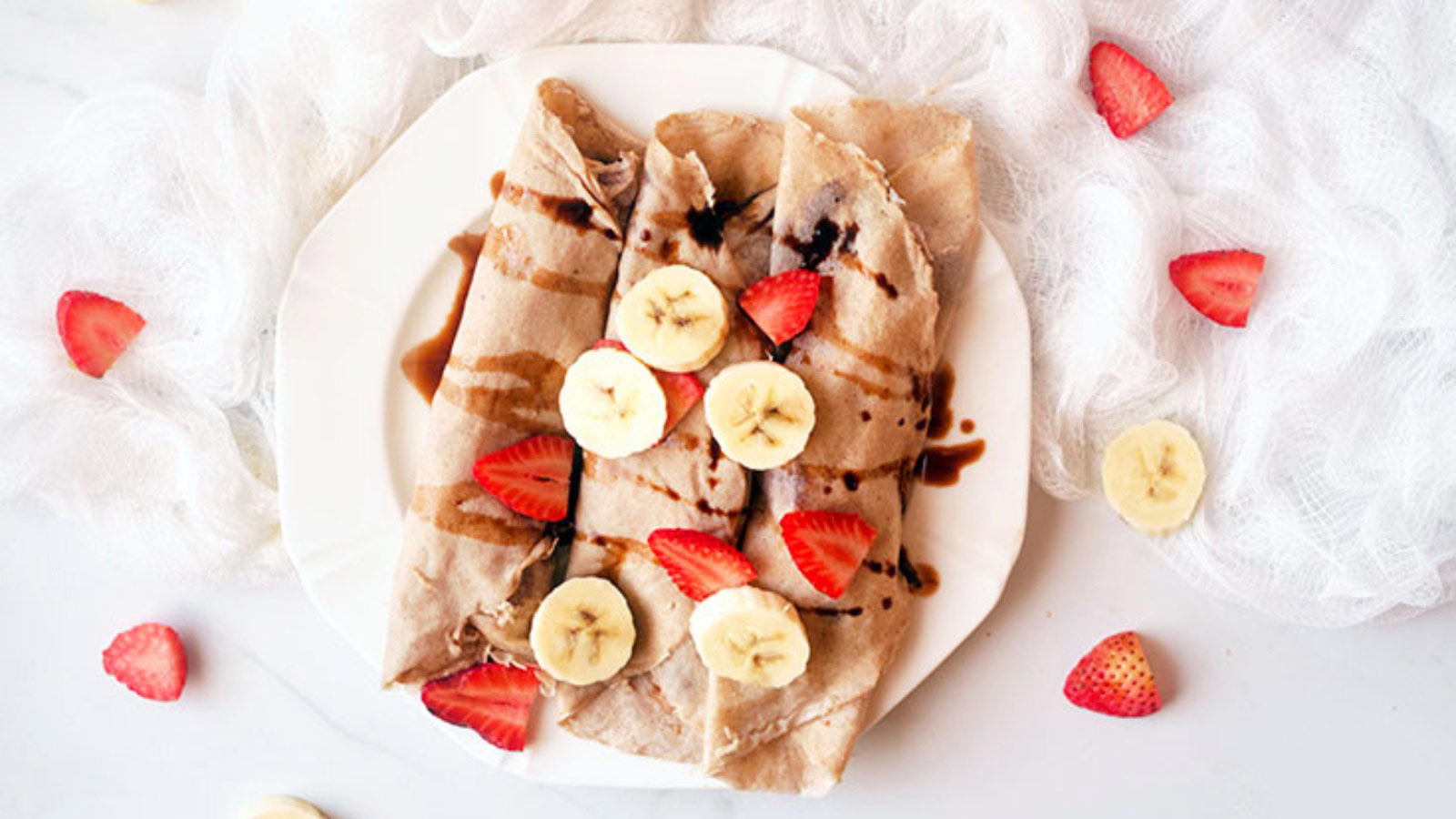 An overhead shot looking down onto a plate of rolled crepes, topped with sliced strawberries and bananas.