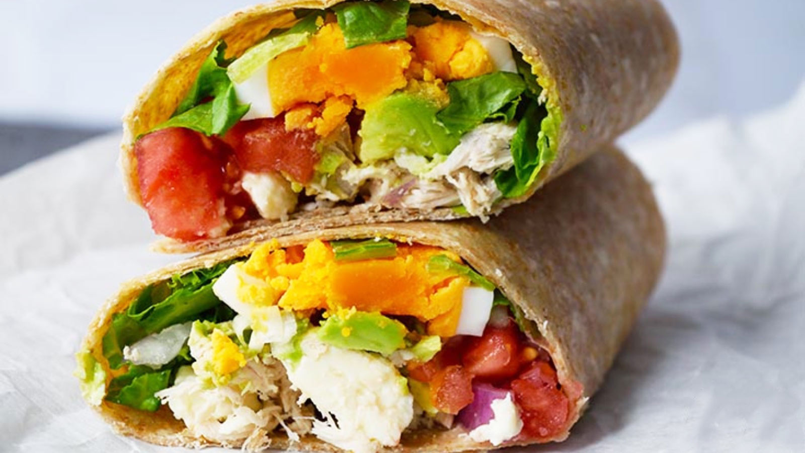 A Cobb Salad Wrap cut in half. The halves are stacked on top of each other on a white piece of parchment paper.