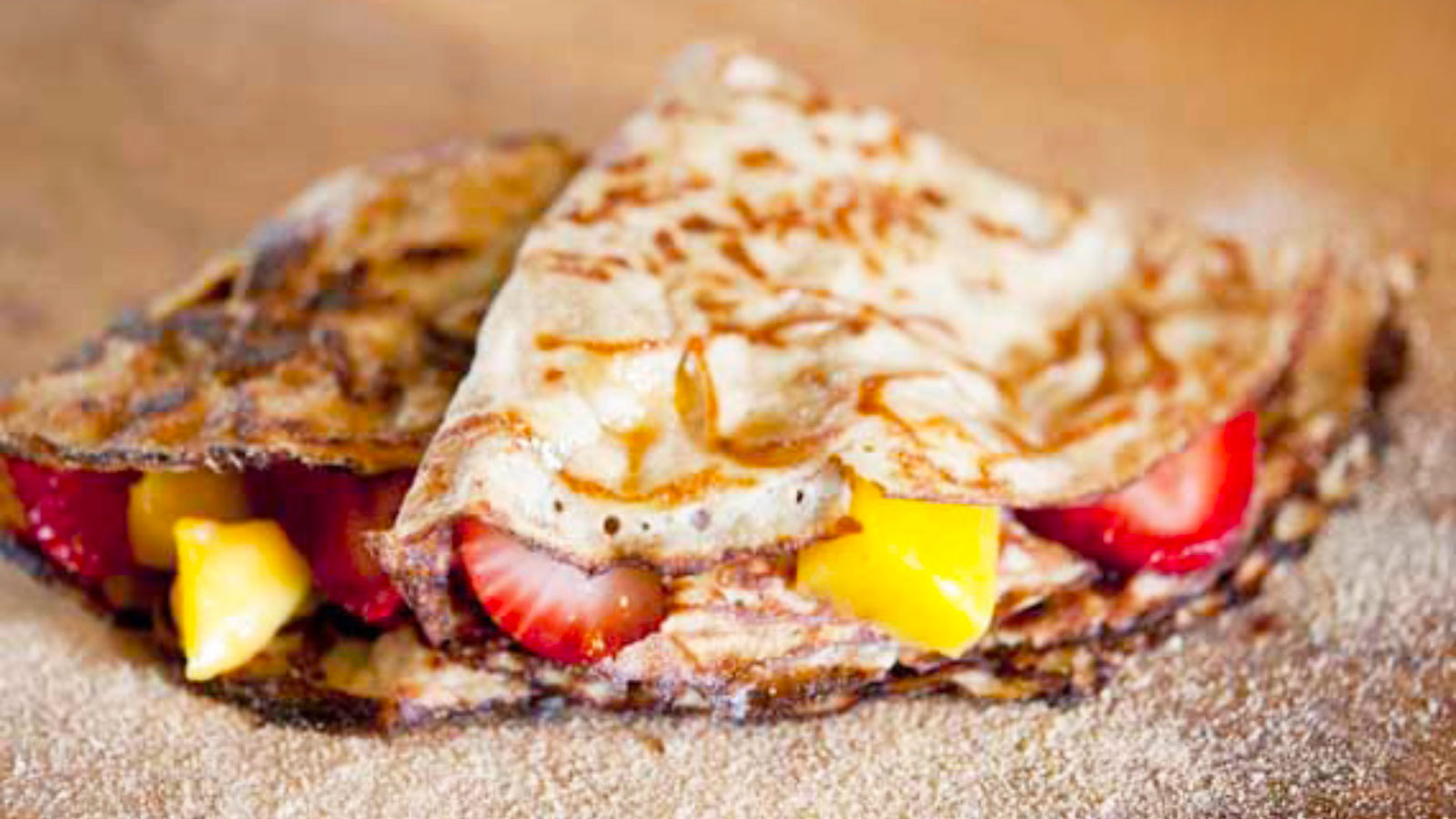 A closeup of two folded crepes stuffed with strawberries and mango and sprinkled with powdered sugar on a wood surface.