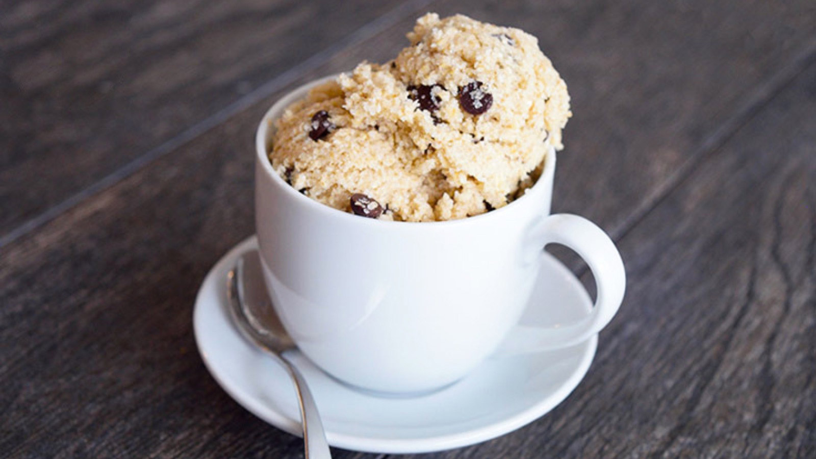 Edible cookie dough served in a white cappuccino mug on a white plate with a spoon.