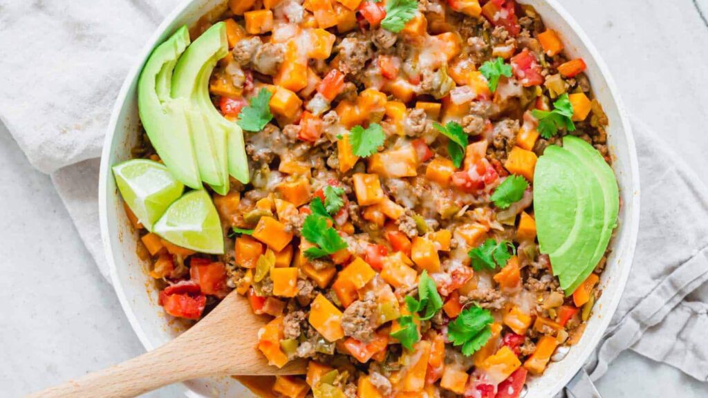 A white bowl filled with ground beef sweet potato skillet and garnished with avocado slices and lime wedges. Overhead focus.