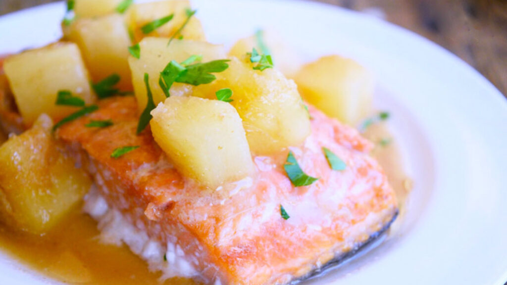 A piece of salmon with pineapple chunks and fresh herbs sprinkled on top sit on a white plate.