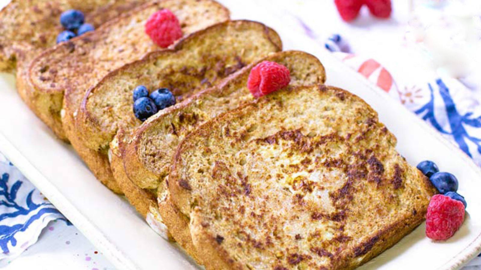 French toast slices lined up on a white platter, sprinkled with a few fresh blueberries and raspberries.