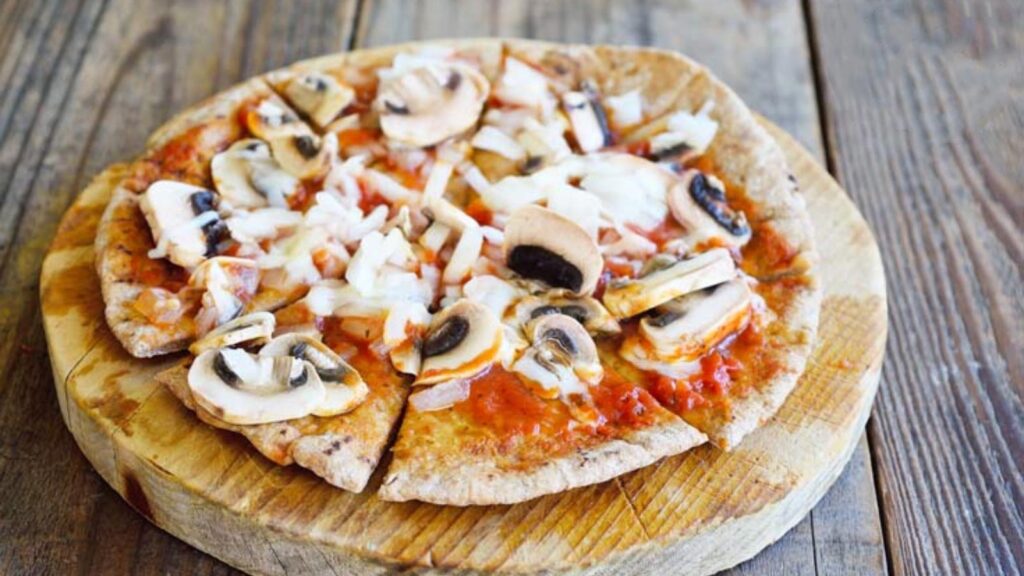 A pita sits on a round cutting board topped with mushrooms, red sauce and melted cheese.