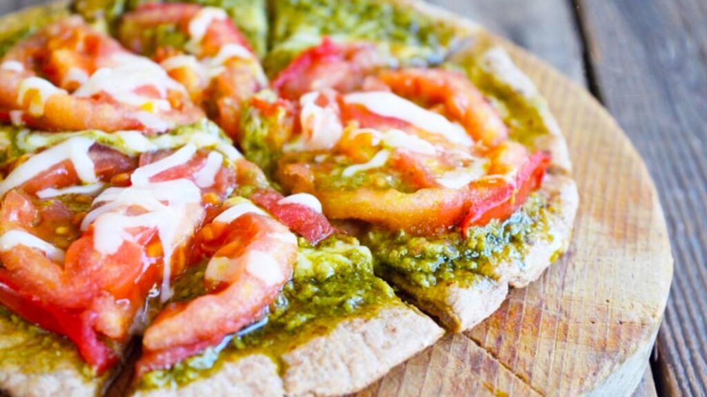 A pita sits on a small round cutting board covered in pesto sauce, fresh tomato slices and melted cheese.