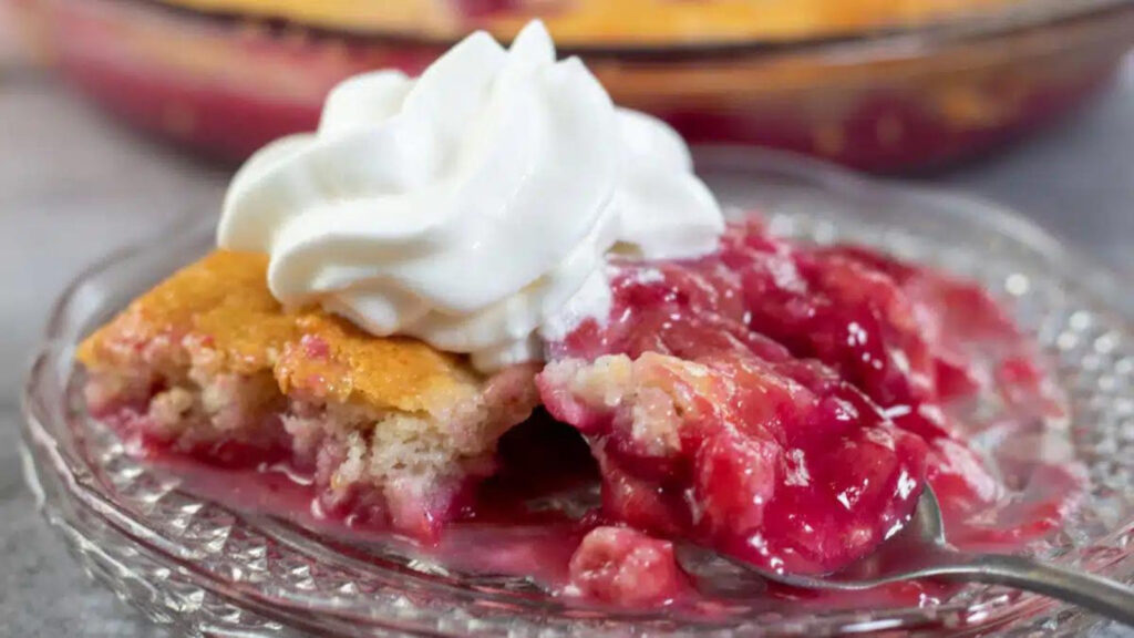 A closeup of a serving of plum cobbler on a glass plate, topped with a dollop of whipped cream.