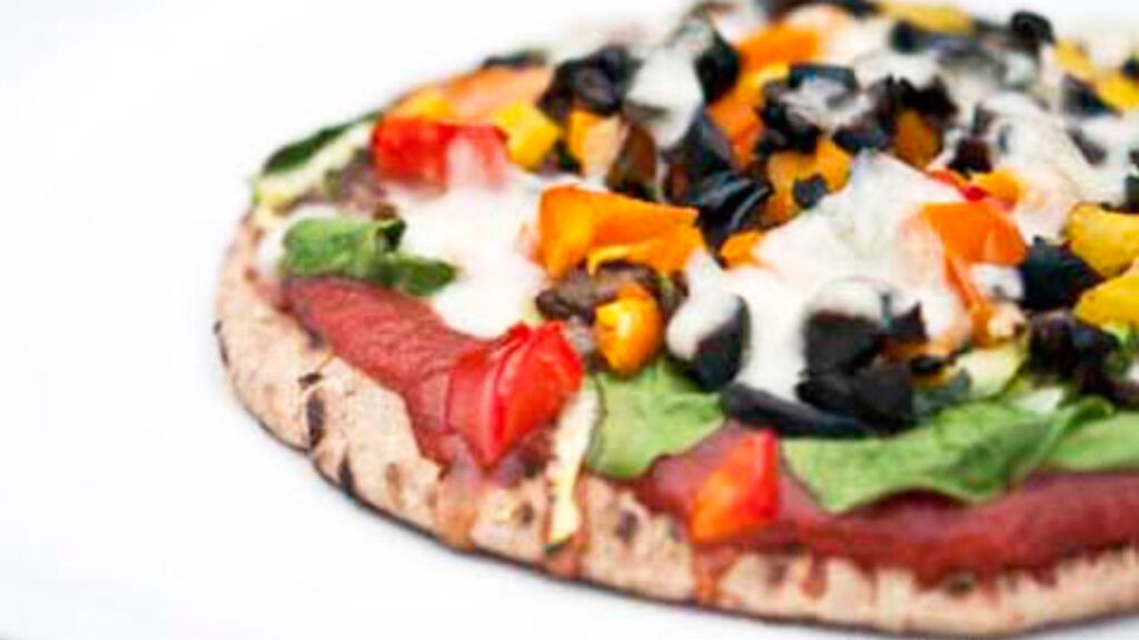 A pita pizza covered in olives, peppers, spinach and cheese sits on a white surface.