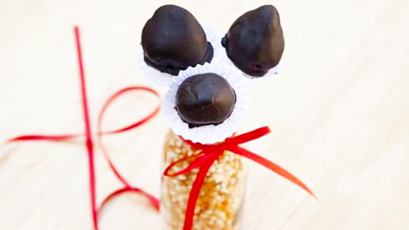 A jar filled with popcorn and tied with a red ribbon holds three Healthy Chocolate Covered Strawberries on sticks.