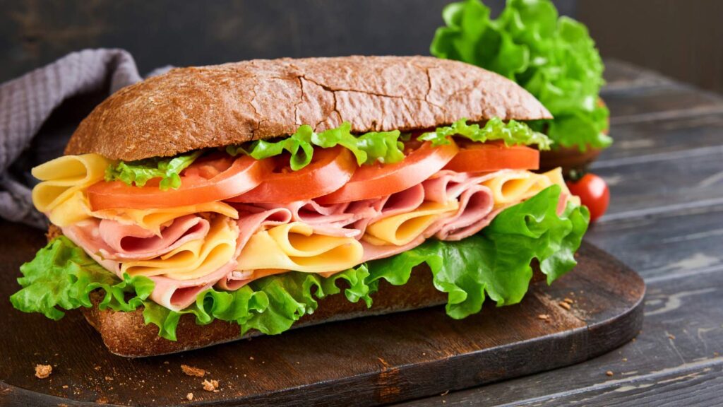 Sandwich. One fresh big submarine sandwich with ham, cheese, lettuce, tomatoes and microgreens on old wooden dark background. Healthy concept, school lunch, breakfast or snack.