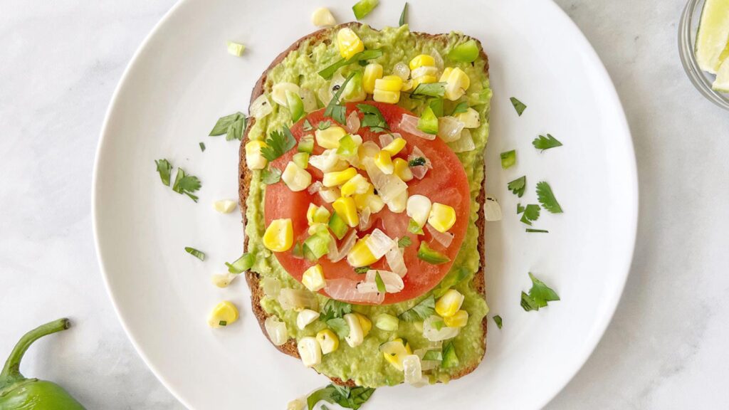 An overhead view of a slice of avocado toast with a fresh tomato slaice and corn salsa on a white plate.