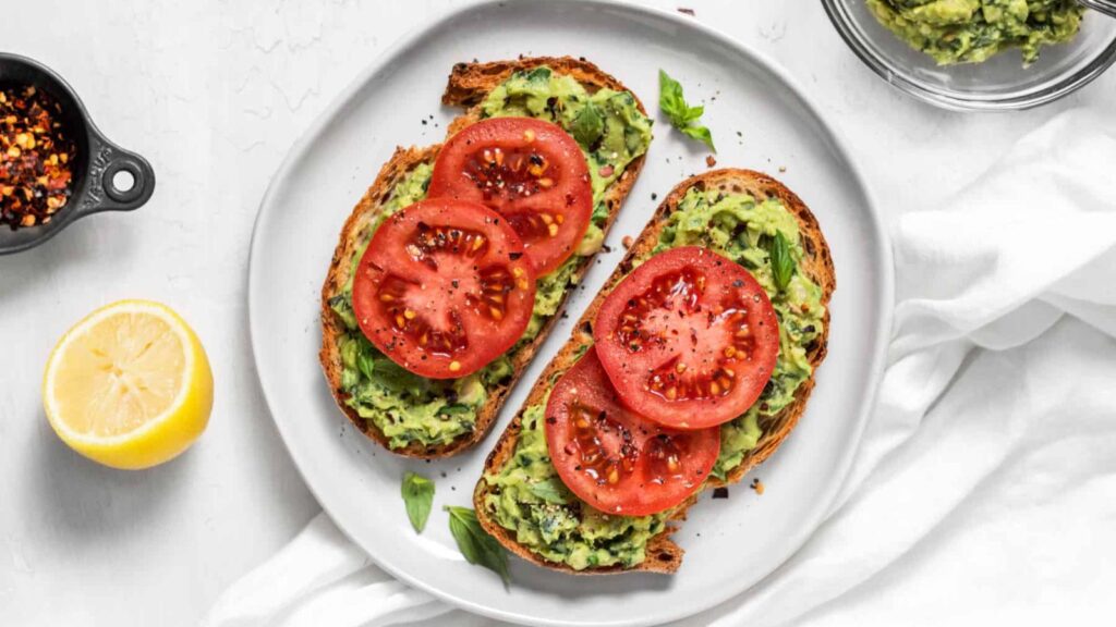 An overhead view of two slices of white bean avocado toast with tomato slices on a white plate.