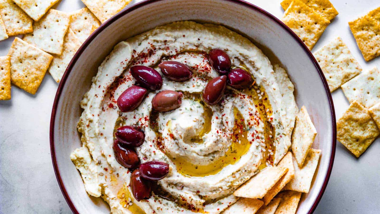 A bowl of hummus with olives and crackers in it.