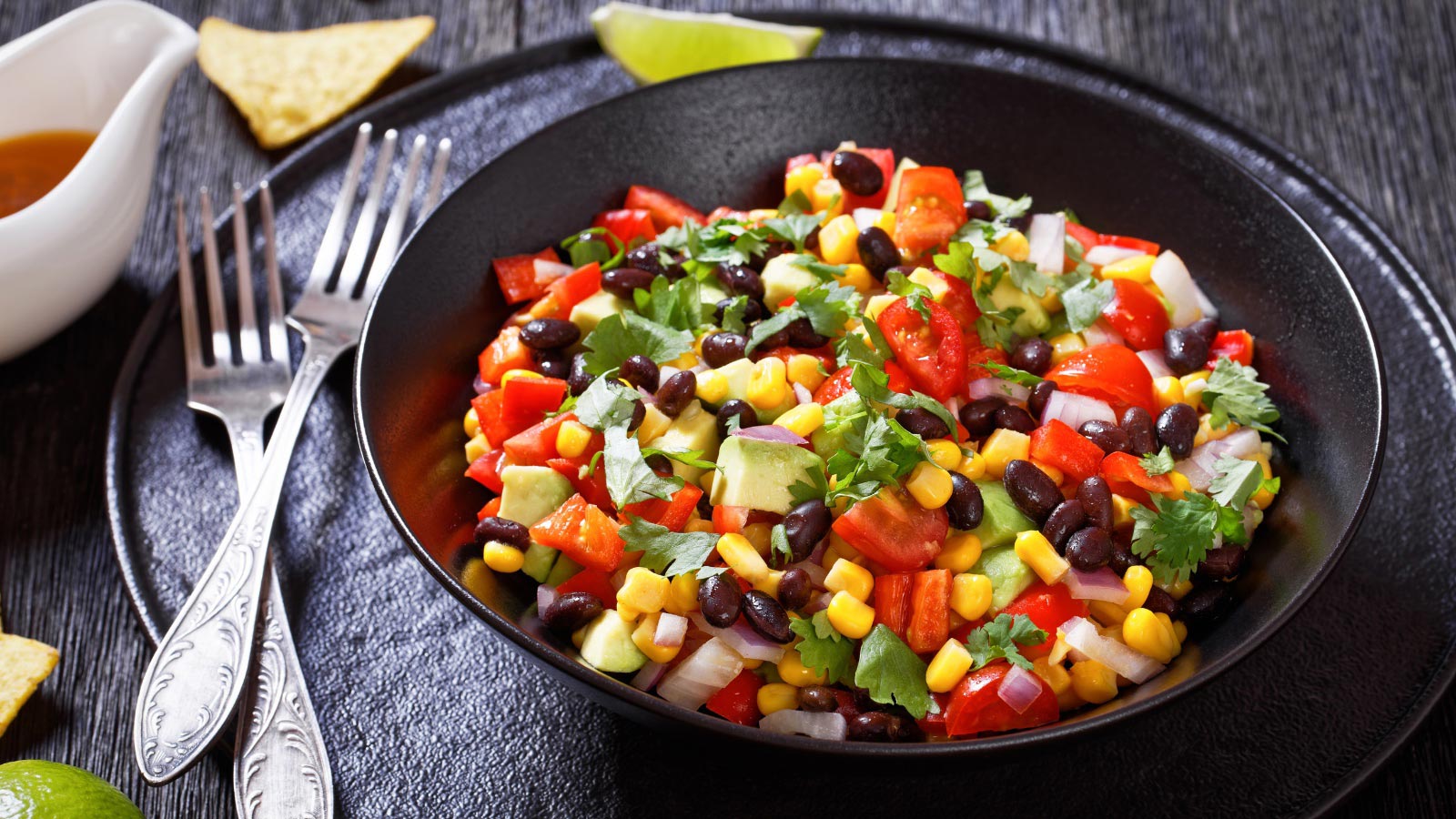 Corn salad, texas caviar with black bean, tomatoes, avocado, red bell pepper, corn, coriander in black bowl on dark wooden table, horizontal view.