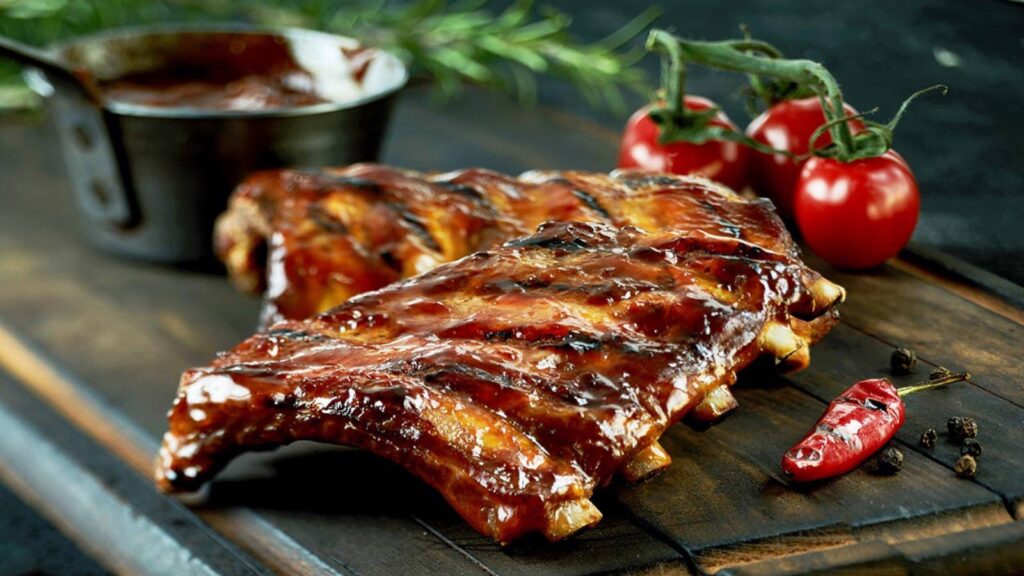 Spicy hot grilled spare ribs from a summer BBQ served with a hot chili pepper and fresh tomatoes on an old vintage wooden cutting board.