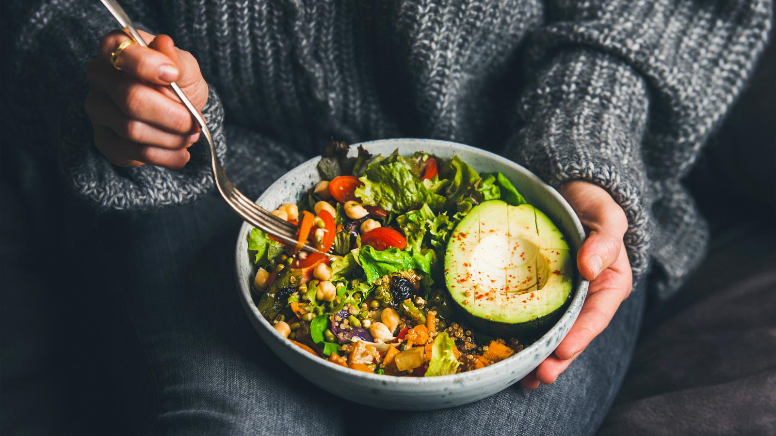 Healthy vegetarian dinner. Woman in jeans and warm sweater holding bowl with fresh salad, avocado, grains, beans, roasted vegetables, close-up.